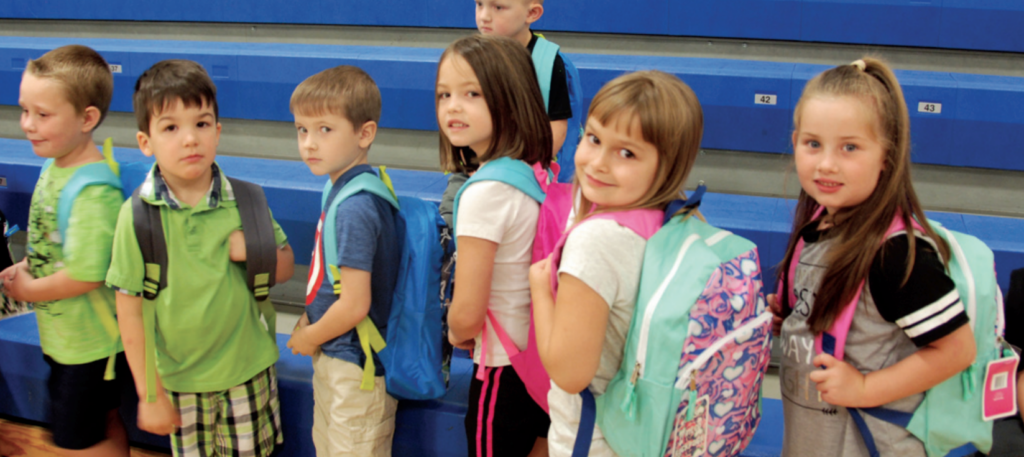 A group of children with backpacks standing in a gymnasium happy through our outreach program.