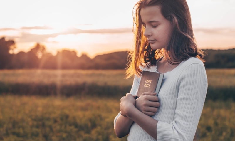 A young girl holding a bible in a field at sunset.