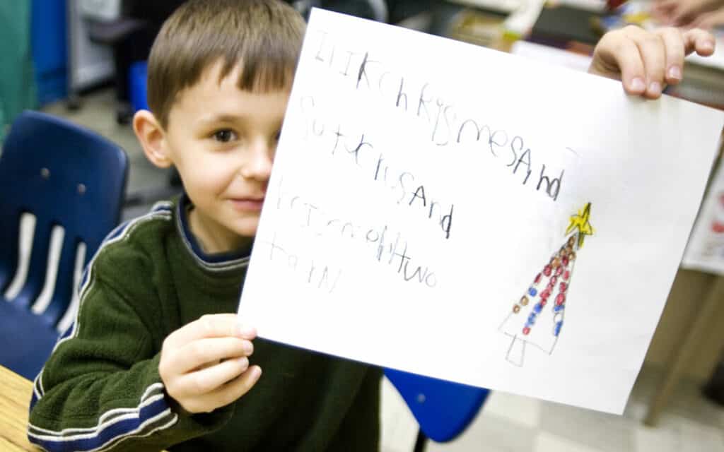 A young boy holding up a paper with a drawing of a christmas tree.