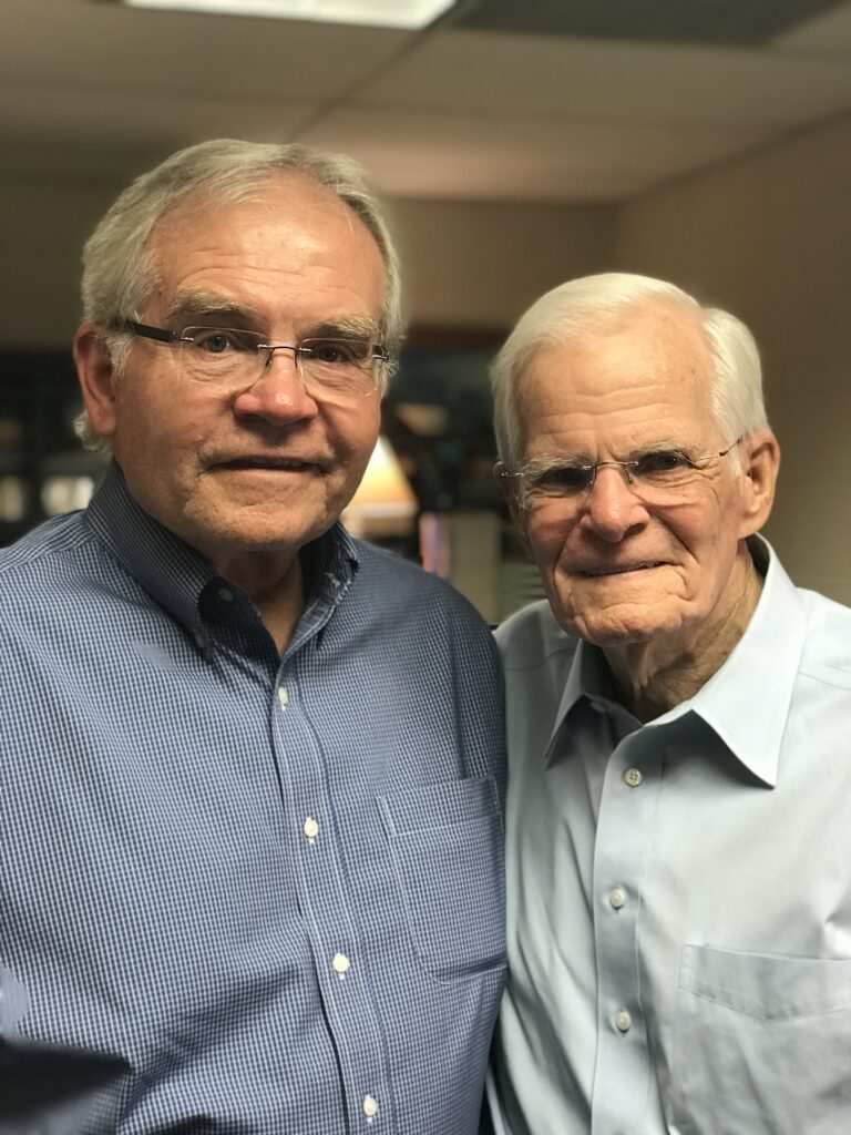 Two elderly men smiling at the camera in an indoor setting.