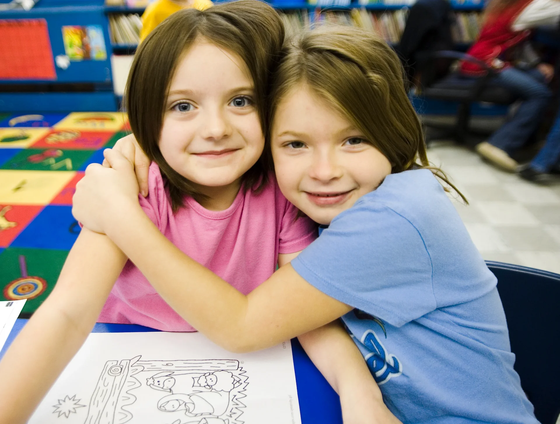 Two young girls embracing at a table in a classroom as part of the back-to-school program.