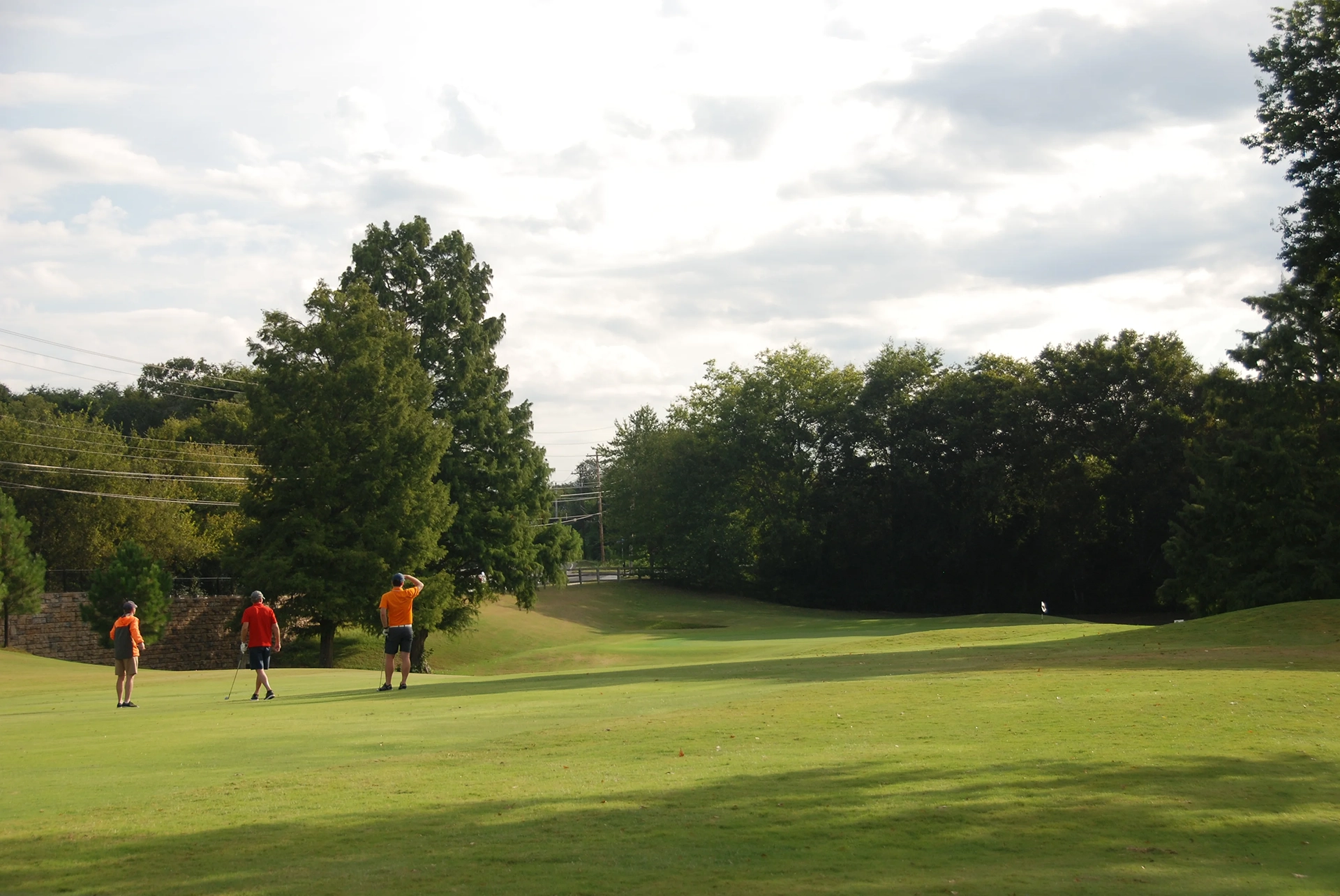 Three people are standing on a golf course, with two holding clubs and one appearing to take a swing. The background includes trees and a partly cloudy sky.